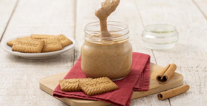 Foto da receita de Pasta de Tostines Especiarias. Observa-se um pote de vidro com a pasta e uma colher mostrando a cremosidade. Biscoitos TOSTINES e canelas em pau decoram a foto.