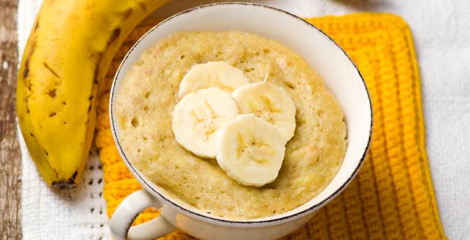 Fotografia em tons de amarelo com uma xícara de café ao centro. Dentro da xícara existe uma receita de bolo de banana. Ao lado existe uma banana inteira em cima de um pano amarelo