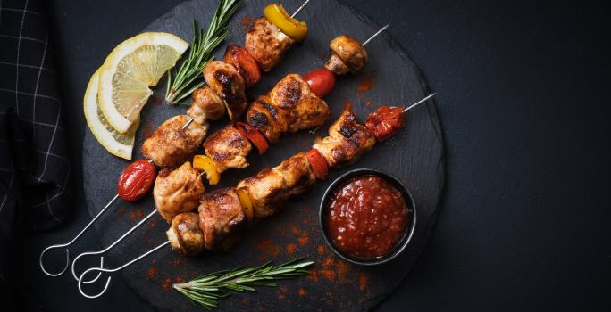 Fotografia vista de cima de três espetinhos, um do lado do outro, de carnes intercaladas com legumes. Os espetos estão sobre um apoio preto redondo, ao lado de duas fatias de limão siciliano, um molho vermelho em um recipiente menor preto e alecrim.