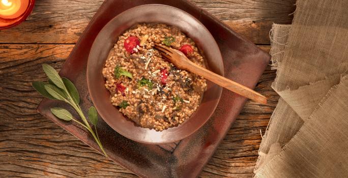 Foto em tons de marrom da receita de risoto ao funghi com aveia corte rústico servida em um prato fundo marrom sobre uma base retangular de mesma cor em cima de uma mesa de madeira