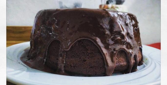 Fotografia em tons de preto em uma mesa de madeira, um pano vermelho, um prato branco raso redondo com o bolo de chocolate com cobertura de chocolate em cima.