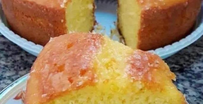 Fotografia de um pedaço grande de bolo de fubá sobre um prato pequeno branco, um pouco fundo, com desenhos delicados. No fundo tem o restante do bolo, quase inteiro, sobre um apoio de plástico branco.