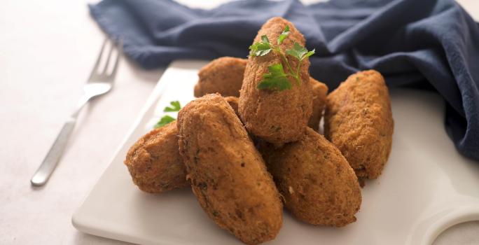 Fotografia de vários bolinhos de bacalhau fritos sobre um apoio branco, ao lado de um pano azul de tom escuro e um garfo.