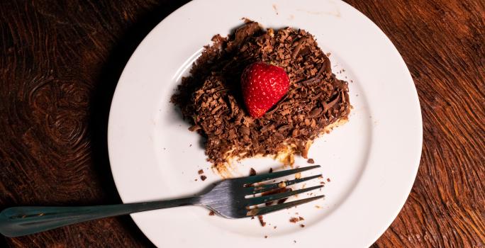 Fotografia vista de cima de um pedaço de pavê com mousse e chocotrio. Por cima tem ganache, raspas de chocolate e morango. O pavê está sobre um prato branco de vidro com um garfo.