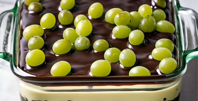 A fotografia mostra um refratário com camadas de uvas verdes sem sementes, cobertas por um creme branco espesso e finalizadas com uma ganache de chocolate ao leite brilhante.