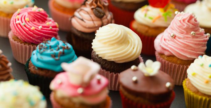 Fotografia de várias unidades de forminhas com cupcakes diversos coloridos e confeitados sobre uma mesa preta.