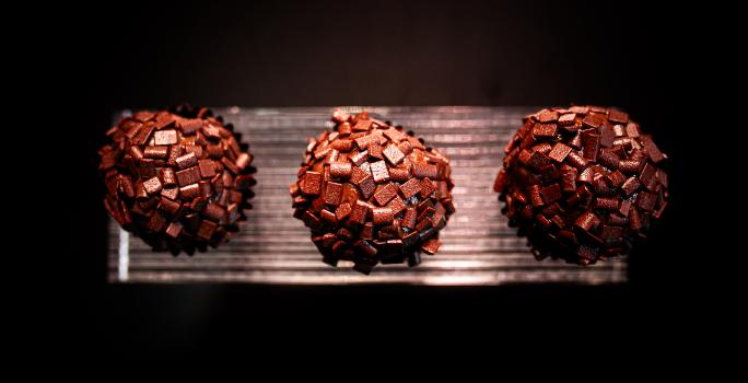 Fotografia vista de cima de três brigadeiros de tucupi reduzido recheados com cupuaçu e enrolados no chocolate amargo. Os brigadeiros estão em um apoio na cor prata, com o fundo escuro.