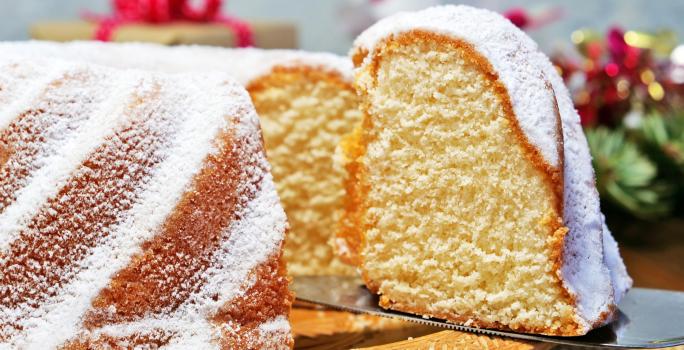 Fotografia de um bolo de massa branca de leite com um pedaço em destaque e cobertura de açúcar de confeiteiro, sobre um apoio reto de tom claro.