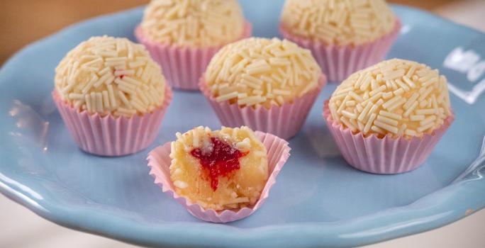 Fotografia de seis forminhas rosa com brigadeiro branco, passado no granulado branco, com recheio de frutas vermelhas. Os brigadeiros estão apoiados em um apoio azul. O brigadeiro da frente está mordido, mostrando a massa e o recheio.