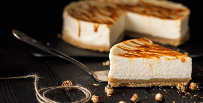 Fotografia de uma fatia de cheesecake com caramelo salgado por cima de uma espátula de prata, sobre uma mesa preta. No fundo tem o resto da cheesecake sobre um apoio preto.