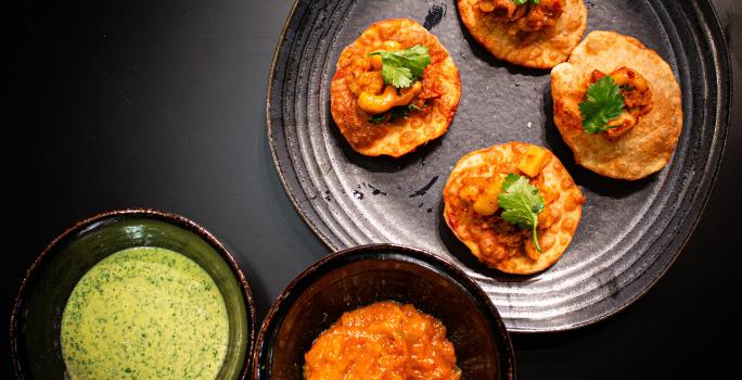 Fotografia vista de cima de um prato preto raso com quatro unidades de pani puri com grão de bico e coentro. Ao lado tem dois molhos em recipientes diferentes, um molho de iogurte e o outro, um molho de manga. Tudo está sobre uma mesa preta.