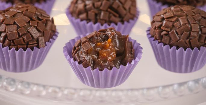 Fotografia em tons de roxo com várias forminhas de brigadeiro roxas ao centro. Dentro de cada forminha existe um brigadeiro de chocolate meio amargo recheado com um creme de caramelo salgado.