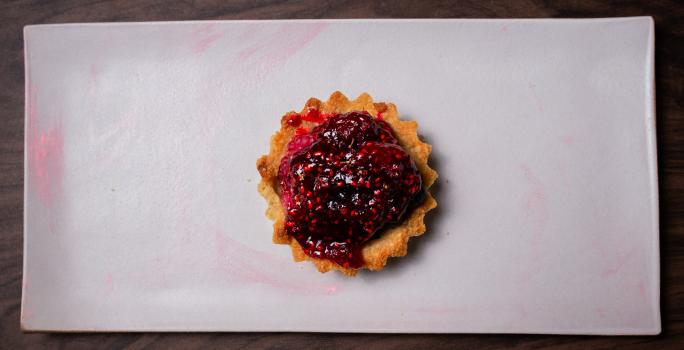 Fotografia em tons de branco com um prato branco ao centro. Em cima do prato existe uma receita de Tartallete coberto com uma geleia de frutas vermelhas e pedaços de frutas vermelhas.