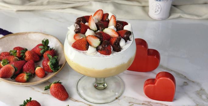 Foto da receita de Morangoffee na Taça, servida em uma grande taça de vidro, em duas camadas, uma de creme bege e outra de creme branco, decorada com morangos com chocolate e suspiros, em uma bancada com mais morangos e alguns corações decorativos