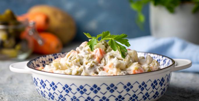 Fotografia de uma salada de maionese que está em um recipiente fundo com desenhos azuis. Tem um ramo de salsinha apoiado na salada. O recipiente está apoiado em uma bancada de textura parecida com gesso, ao fundo tem frutas.