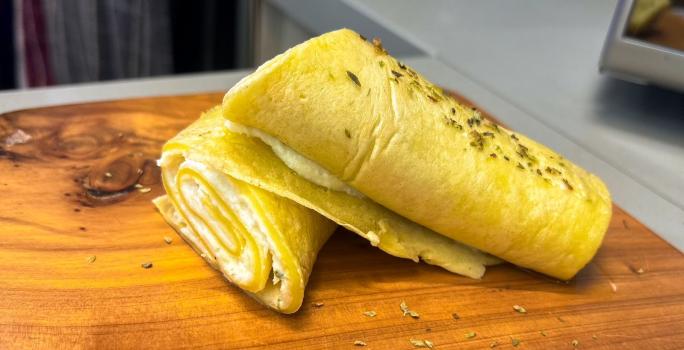 Fotografia em tons de amarelo com uma tábua de madeira ao centro. Em cima da tábua existe uma crepioca recheada com creme de ricota e Ninho Adulto