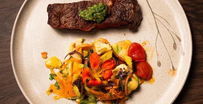 Fotografia vista de cima de um bife ancho com pesto de cerefólio e salada de cogumelos ao lado. A receita está em um prato grande, branco e raso, o qual está sobre uma mesa de madeira.