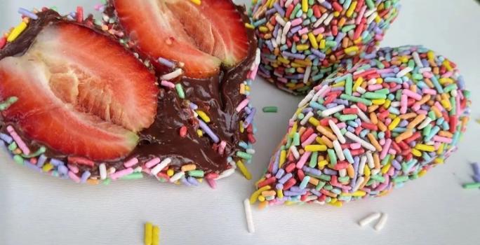 Fotografia em tons coloridos em uma bancada de madeira branca. Ao centro, há um morango cortado ao meio e ao lado, há 2 coxinhas de brigadeiro e uma colher de brigadeiro.