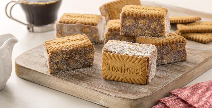 Fotografia de uma tábua de madeira com sete unidades de palha italiana com tostines especiais, ao lado de três biscoitos tostines. No fundo tem uma xícara de vidro com café.