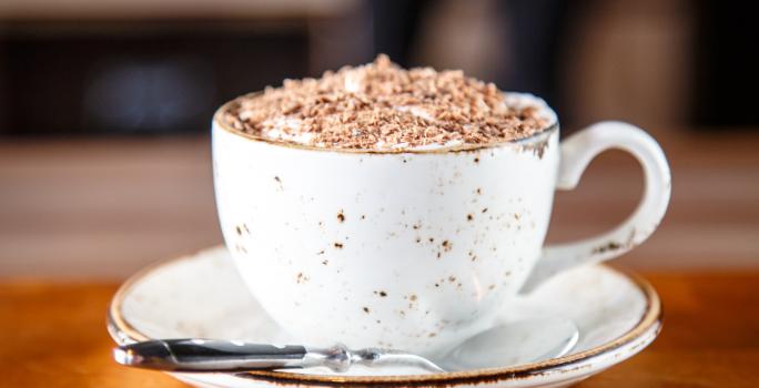 fotografia em tons de branco e marrom de uma bancada marrom vista de frente. Ao centro contém um pratinho redondo branco com uma xicara branca por cima, dentro da xicara contém a bebida e por cima raspas de chocolate, ao lado uma colher para mexer.