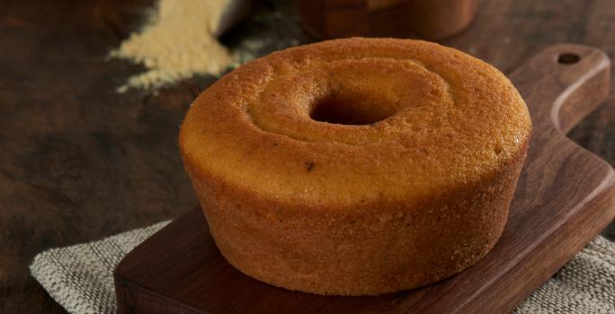 Foto aproximada do bolo de milho com erva-doce, redondo, com furo central, bem dourado, sobre uma tábua de madeira com um punhado de fubá aparecendo ao fundo