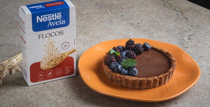 Foto da receita de Torta de Maçã com Chocolate e Nestlé Aveia Flocos, decorada com mirtilos, amoras e hortelã, servida em um prato laranja, sobre uma bancada de pedra onde está uma caixa de Nestlé Aveia Flocos e um ramo de trigo seco