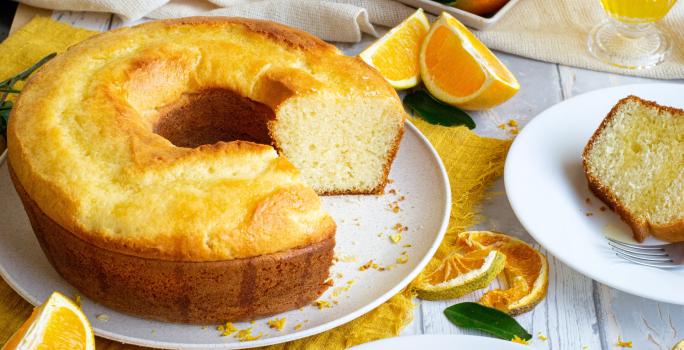 Fotografia de um bolo de laranja feito em forma de furo central sobre um prato raso de cerâmica branco. Ao lado tem um prato de sobremesa com uma fatia do bolo e um garfo, e ao redor tem pedaços e fatias de laranja, sobre uma mesa de madeira branca.