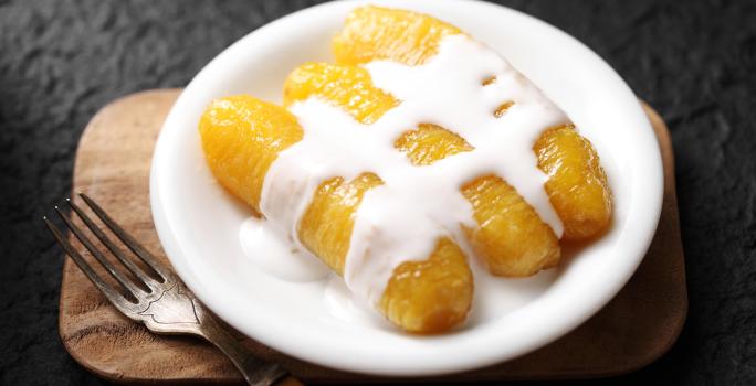 Fotografia de três bananas carameladas com creme de leite por cima.  As bananas estão em um prato pequeno branco raso, ao lado de um garfo que está apoiado em uma tábua de madeira pequena.