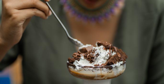 Fotografia de uma pessoa segurando uma colher de sopa e comendo uma sobremesa de camadas de massa e MOÇA Leite Condensado Cremoso. O doce está em uma tacinha de vidro.