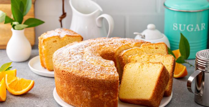 Fotografia de um bolo de laranja feito em forma redonda com furo no meio, o bolo está sobre um apoio, e tem três fatias cortadas, uma apoiada na outra. Ao fundo tem uma fatia do bolo em um prato de vidro pequeno, ao lado de pedaços de laranja.