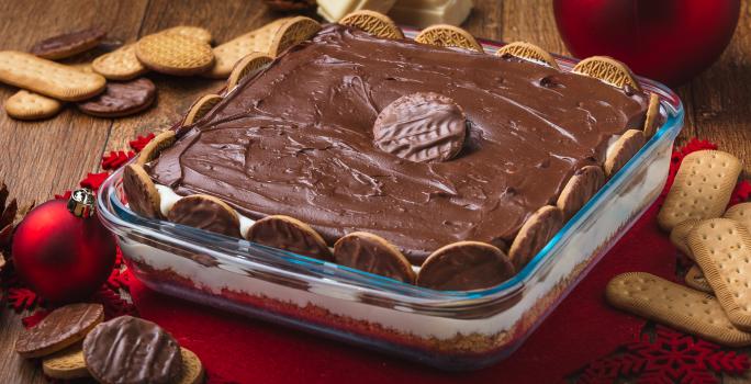 Foto da receita de Torta Holandesa na Travessa, em uma bancada de madeira decorada com temas de natal, alguns biscoitos espalhados, pedaços de chocolate branco. A receita tem um creme marrom sobre um creme claro e está em um refratário de vidro