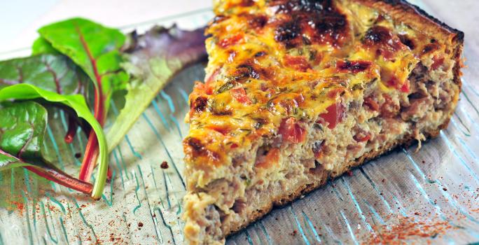 Fotografia mostra uma fatia de torta com camadas bem definidas de massa leve e recheio colorido de sardinha, tomate e azeitonas, polvilhada com orégano.