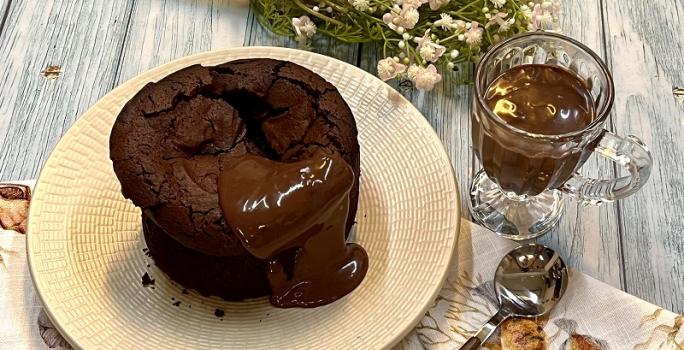 Foto em tons de bege e marrom da receita de bolo fudge servida sobre um prato bege e uma tolha de mesa decorada. Ao lado, há uma colher prateada e uma caneca de vidro com chocolate derretido. Ao fundo, um buqué de flores cor de rosa