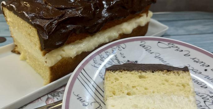 Foto da receita de Boston Pie. Observa-se o bolo retangular com uma camada de creme branco no meio e cobertura de chocolate no plano de fundo. À frente, um prato de sobremesa redondo com uma fatia do bolo.