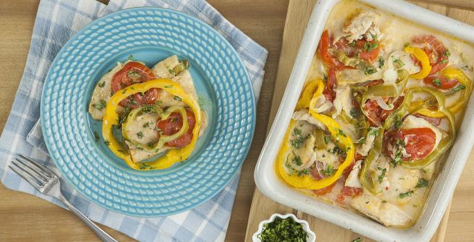 Fotografia em tons de azul em uma bancada de madeira, um pano azul xadrez, um prato azul com um pouco da moqueca de peixe com pimentões vermelho e amarelo, uma tábua de madeira e em cima dela uma travessa com a moqueca com os pimentões.