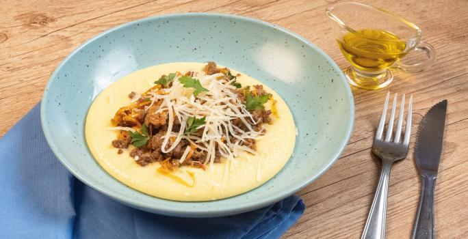 Fotografia de um prato fundo azul claro com uma polenta cremosa, e por cima, uma carne moída com cheiro verde e queijo parmesão ralado. O prato está apoiado em um pano azul pequeno que está sobre uma mesa de madeira, e ao lado tem talheres e azeite.