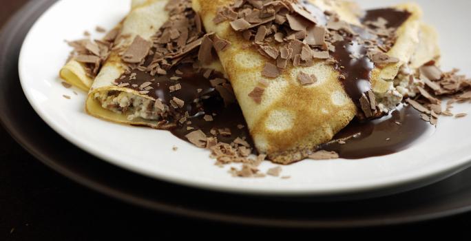 Fotografia de algumas panquecas, uma por cima da outra, recheadas e cobertas de brigadeiro, e por cima, raspas de chocolate. As panquecas estão em um prato branco que está sobre um sousplat marrom escuro.