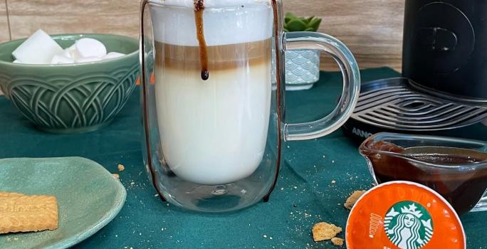 Fotografia em tons de verde em uma bancada com um pano verde, uma xícara de vidro grande com a bebida de leite e café preparada dentro dela. Decorada com marshmallow, calda de chocolate e biscoito triturado.