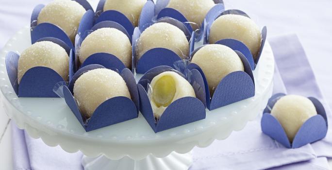 Fotografia em tons de branco e azul de uma bancada branca com paninho azul, sobre ele uma bailarina branca com docinhos de brigadeiro branco com uva verde dentro de forminhas quadradas azuis.