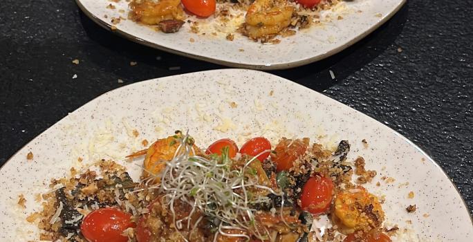 Fotografia de dois pratos de vidro parcialmente quadrados, de pontas arredondadas, sobre uma mesa preta. Dentro dos pratos tem uma massa linguine nero com molho rústico de tomate grape, camarões e farofa de castanhas.