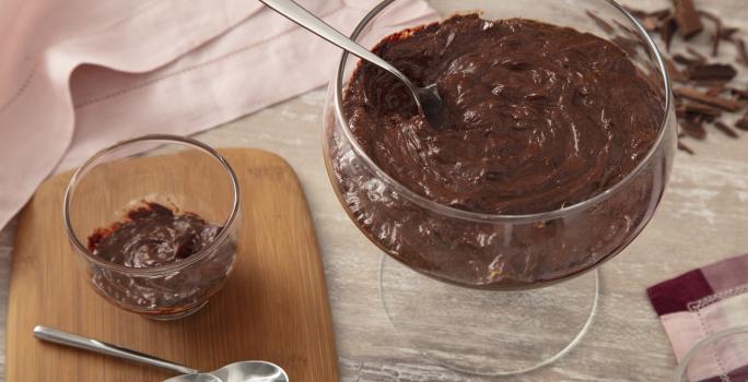 Foto da receita de Mousse Fácil de Chocolate. Observa-se um recipiente redondo com a mousse e, ao lado, um recipiente individual. Também tem duas colherinhas ao lado e raspas de chocolate como decoração.