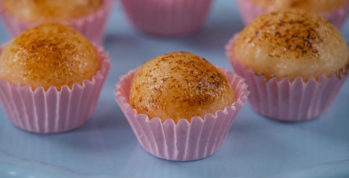 Fotografia de seis unidades de embalagens rosas, uma perto da outra, formando um hexágono. em cada forminha, tem um brigadeiro de creme brulée. As embalagens estão sobre um apoio de cor azul claro.