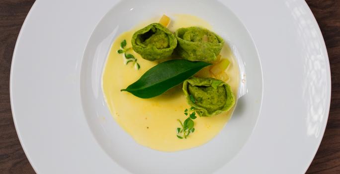 Fotografia em tons de branco com um prato branco ao centro. Em cima do prato existe uma receita de massa recheada de cor verde acompanhada de um molho de cor amarela.