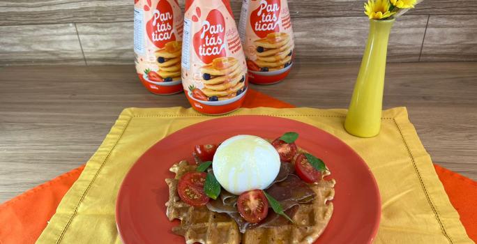 Fotografia em tons de vermelho com um prato vermelho ao centro. Em cima do prato existe uma receita de waffle com burrata, presunto de parma, tomate cereja e manjericão. Ao fundo existe três garrafas de PANTÁSTICA, um produto para fazer panquecas.