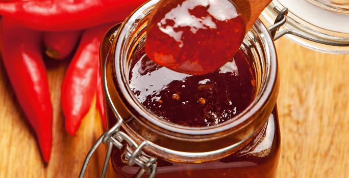 Fotografia de um recipiente de vidro decorativo com geleia de pimenta e laranja. O recipiente está sobre uma mesa de madeira, ao lado de várias pimentas dedo-de-moça.