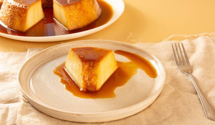 Imagem de um prato claro rústico com uma fatia de pudim de pão e calda de caramelo. Ao fundo, um prato com o restante do pudim. Imagem de um prato claro rústico com uma fatia de pudim de pão e calda de caramelo. Ao fundo, um prato com o restante do pudim.